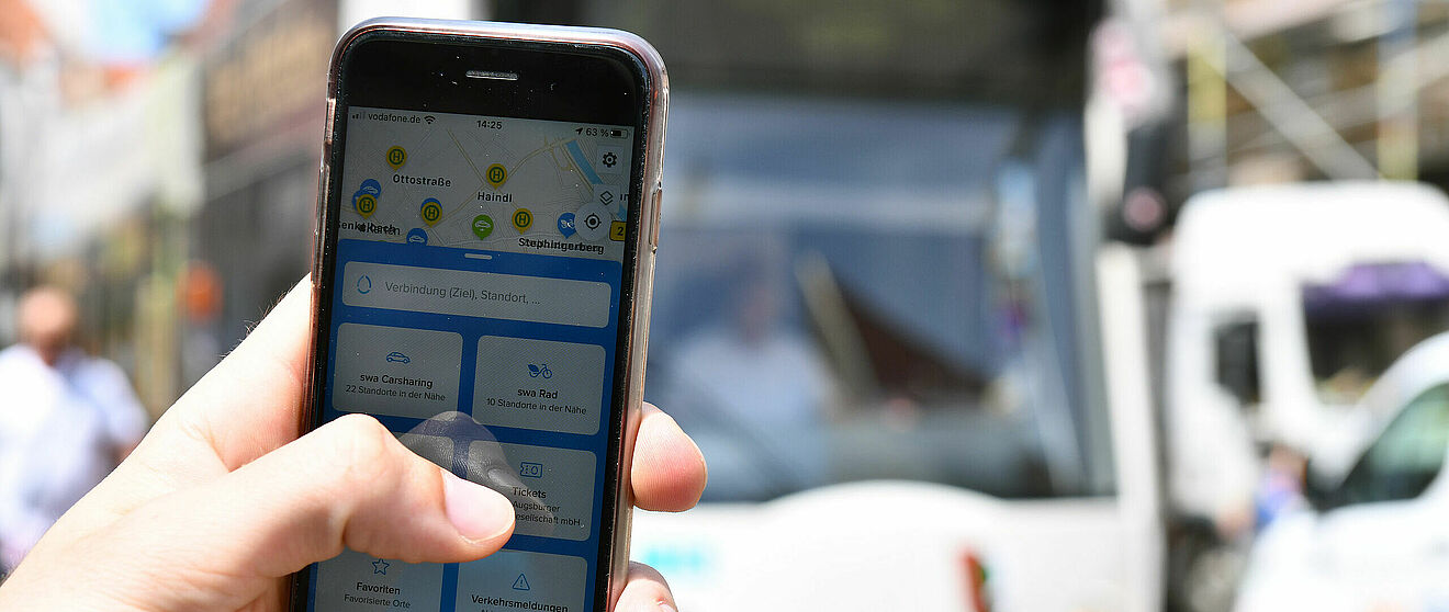 Nahaufnahme von einer Hand, welche ein Handy mit geöffneter swa Mobil-App hält. im Hintergrund eine swa Straßenbahn.