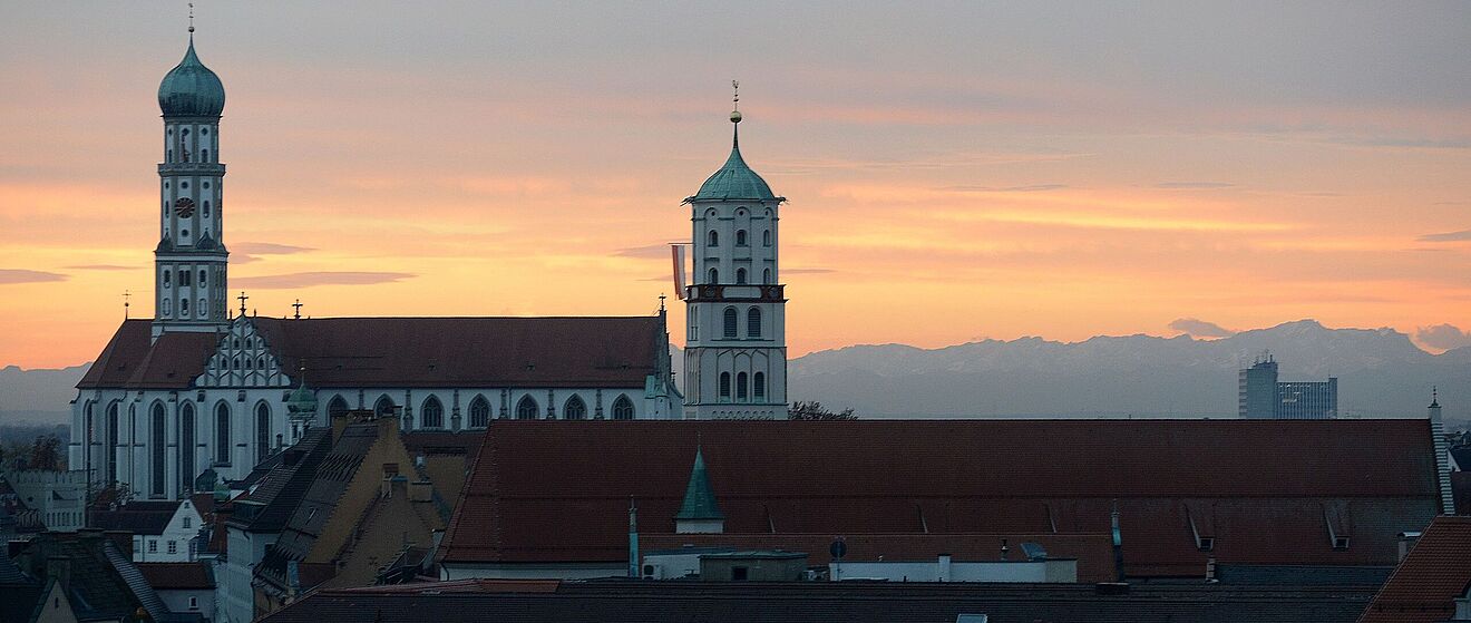 Eine Abendaufnahme zeigt die Basilika St. Ulrich und Afra in Augsburg, deren charakteristische Türme sich gegen einen orange-gelb gefärbten Himmel bei Sonnenuntergang abheben.