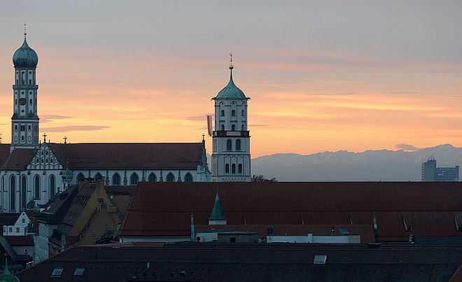 Eine Abendaufnahme zeigt die Basilika St. Ulrich und Afra in Augsburg, deren charakteristische Türme sich gegen einen orange-gelb gefärbten Himmel bei Sonnenuntergang abheben.