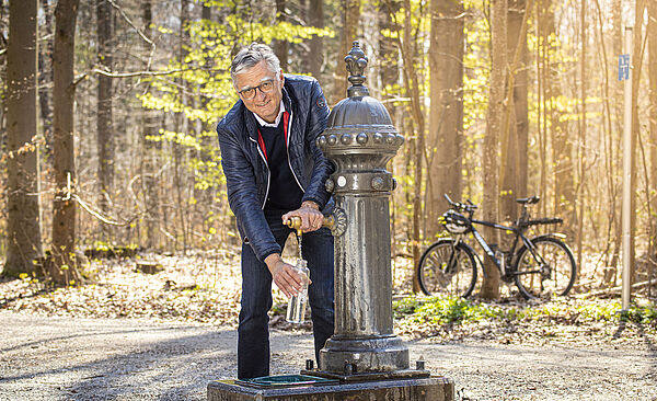 Älterer Mann mit Brille füllt eine Flasche mit Wasser aus einem Trinkwasserbrunnen im Wald auf. Im Hintergrund ist ein Fahrrad.