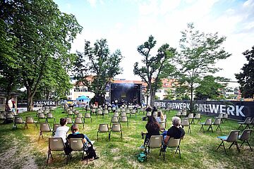 Menschen sitzen auf Stühlen auf dem Gaswerksgelände im Hintergrund ist eine Bühne mit Musikern