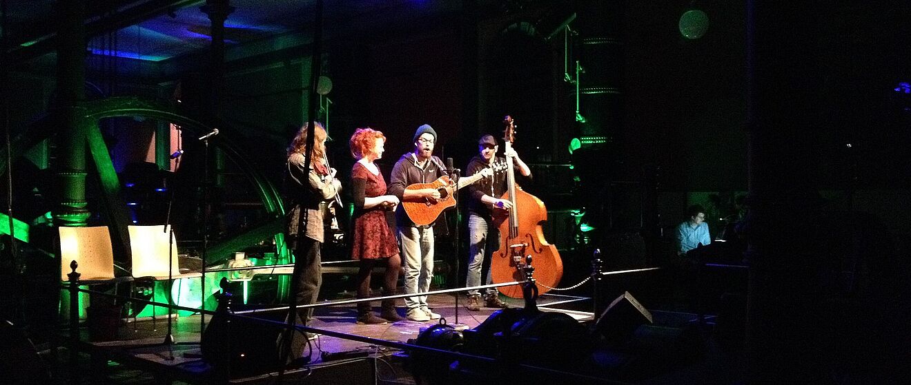 Zwei Frauen und zwei Männer stehen mit Geige Gitarre und Kontrabass auf einer Bühne.