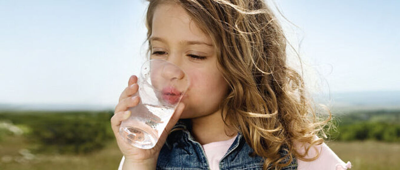 Kleines Mädchen trink Wasser aus einem Glas. Im Hintergrund ist Natur zu sehen.