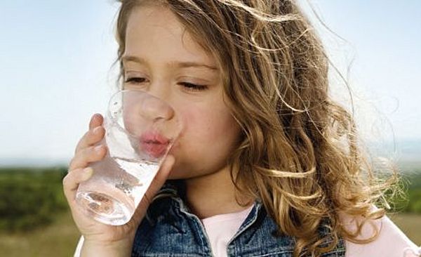 Kleines Mädchen trinkt Wasser aus einem Glas. Im Hintergrund ist Natur zu sehen.
