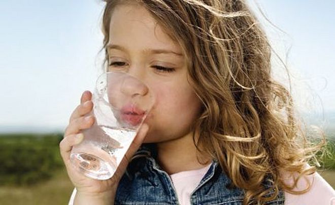 Kleines Mädchen trinkt Wasser aus einem Glas. Im Hintergrund ist Natur zu sehen.