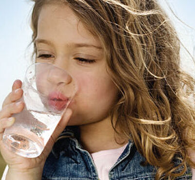 Kleines Mädchen trinkt Wasser aus einem Glas. Im Hintergrund ist Natur zu sehen.