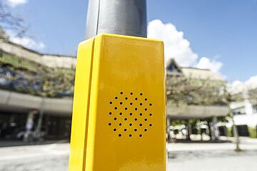 Ein gelbes Kästchen mit einem Lautsprechergitter, das an einem Laternen- oder Ampelmast befestigt ist. Ein klick auf das Bild ermöglicht eine größere Ansicht