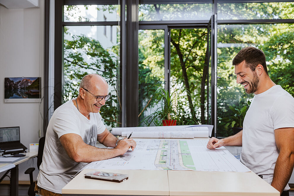 Zwei lachende Bauingenieure vor einem Plan auf dem Tisch