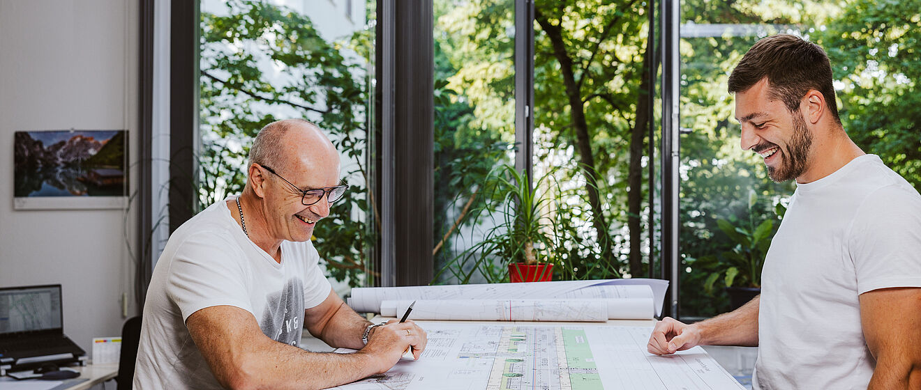 Zwei lachende Bauingenieure vor einem Plan auf dem Tisch