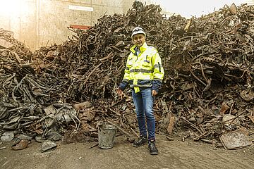 Person mit Helm und neongelber Schutzjacke steht vor einem großen Haufen Rohstoffen aus der Mülltrennung. Ein Klick sorgt für eine Großansicht des Bildes.