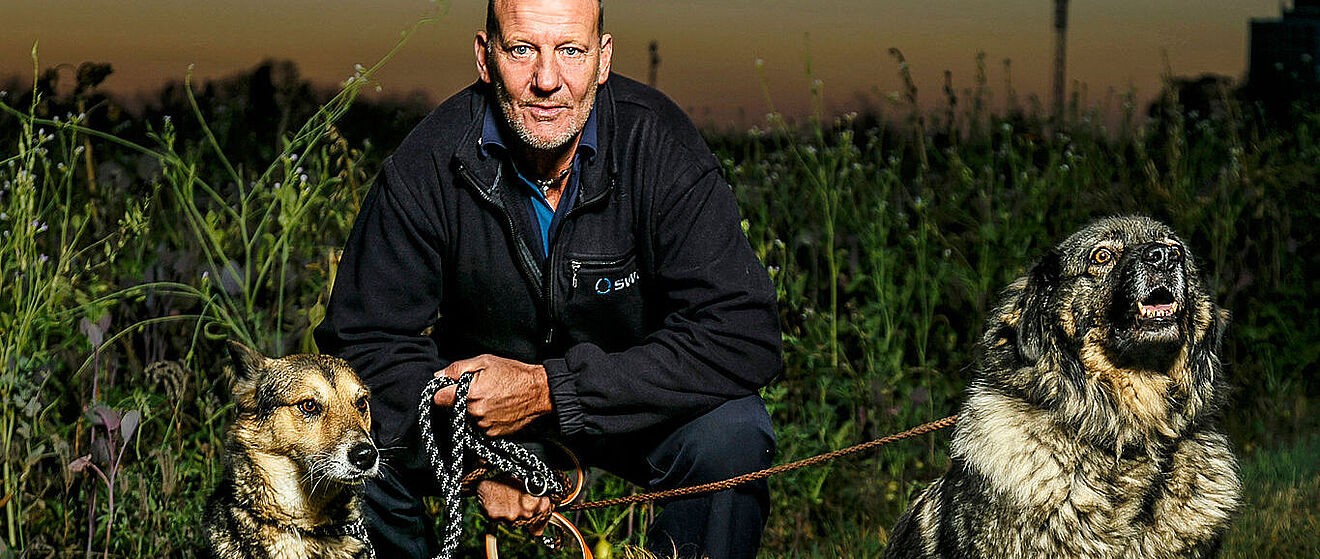 Ein Mann in der Hocke hält auf einer Wiese zwei angeleinte Hunde links und rechts.