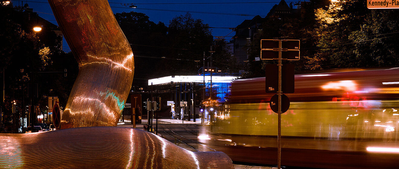 Im Vordergrund befindet sich das Kunstwerk mit dem Namen "Ostern". Dahinter bewegt sich ein Fahrzeug sehr schnell entlang den Gleisen auf dem Boden. Im Hintergrund befindet sich eine der Augsburger Haltestellen.