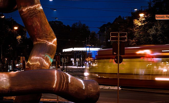 Im Vordergrund befindet sich das Kunstwerk mit dem Namen "Ostern". Dahinter bewegt sich ein Fahrzeug sehr schnell entlang den Gleisen auf dem Boden. Im Hintergrund befindet sich eine der Augsburger Haltestellen.
