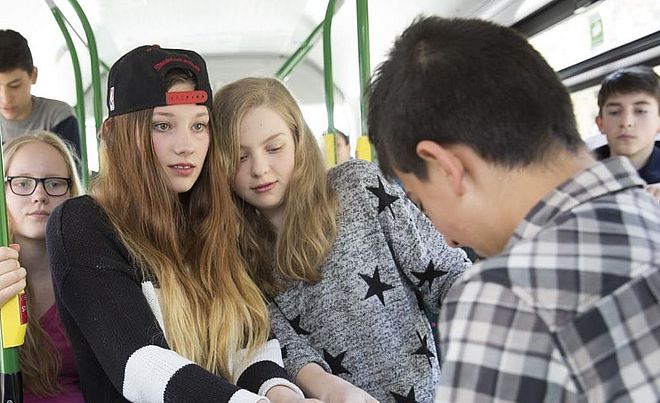 Mehrere Kinder stehen in einem Bus. Im Zentrum ist ein Mädchen mit Kappe, dass den Pulli eines Jungen aus der Hand zieht.