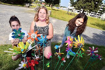 Drei Mädchen stecken gebastelte Blumen in eine Wiese auf dem Gaswerksgelände. - Beim anklicken wird das Bild vergrößert.
