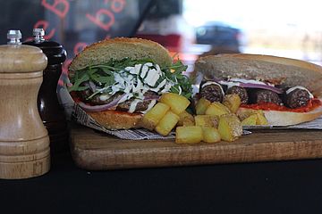 Ein Burger mit Pommes und Cevapcici im Fladenbrot liegen auf einem Holzbrett. Daneben sind Gewürze