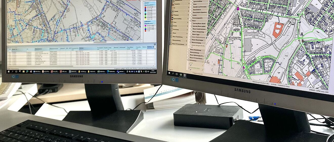 Schreibtisch mit einer Tastatur und zwei Bildschirmen auf denen Stadtpläne sind.