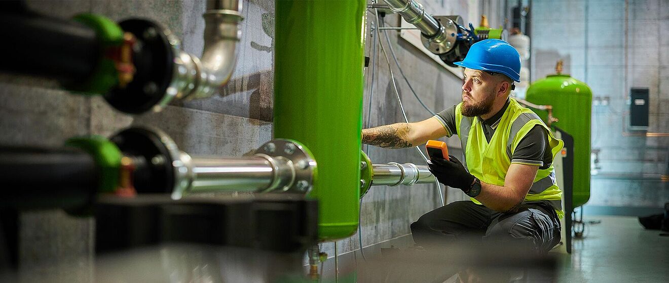 Mann mit Helm, der Anlagen überprüft und ein Messgerät hält.