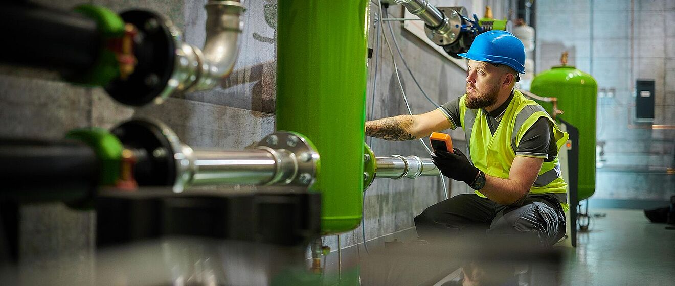 Mann mit Helm, der Anlagen überprüft und ein Messgerät hält.