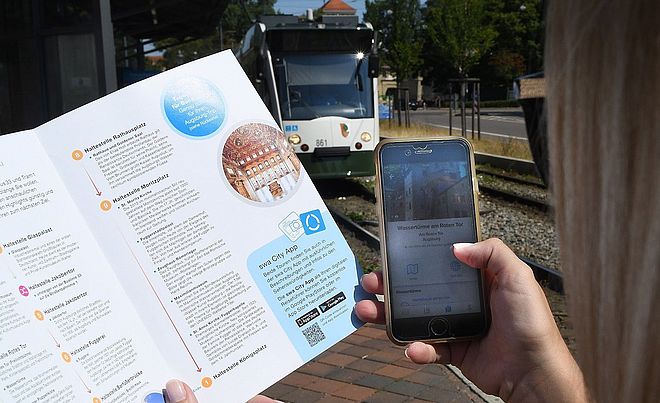 Person hält in der linken Hand ein Programmheft der Entdeckertour und in der rechten Hand ein Smartphone mit der swa City App. Im Hintergrund ist eine Straßenbahn.