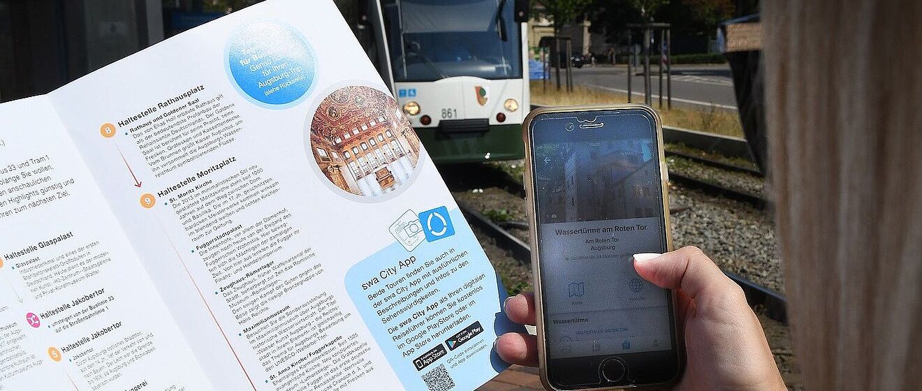 Person hält in der linken Hand ein Programmheft der Entdeckertour und in der rechten Hand ein Smartphone mit der swa City App. Im Hintergrund ist eine Straßenbahn.