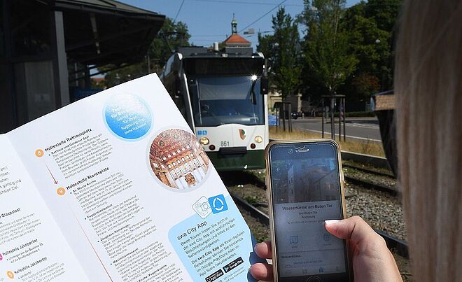 Person hält in der linken Hand ein Programmheft der Entdeckertour und in der rechten Hand ein Smartphone mit der swa City App. Im Hintergrund ist eine Straßenbahn.