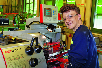Junge steht vor einem Arbeitsbereich mit vielen Knöpfen und Hebeln, er trägt eine Schutzbrille und schaut in die Kamera. - Beim anklicken wird das Bild vergrößert.
