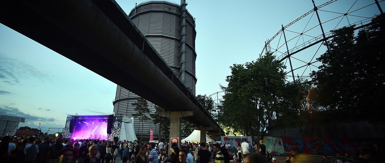 Menschen laufen auf dem Gaswerk im Hintergrund ist eine beleuchtete Konzertbühne und der Gaskessel. Weiterverlinkung auf Festivalsommer Newsartikel