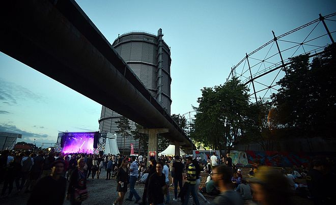 Menschen laufen auf dem Gaswerk im Hintergrund ist eine beleuchtete Konzertbühne und der Gaskessel. Weiterverlinkung auf Festivalsommer Newsartikel