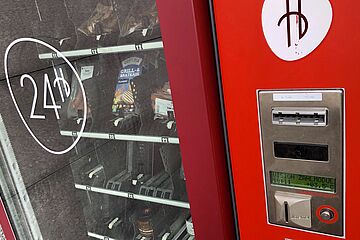 Ein roter Automat mit Münzeinwurf, Display und Tastenfeld, daneben hinter Glas verschiedene Lebensmittel zum Grillen. - Ein Klick auf das Bild führt zur Vergrößerung der Fotoansicht.
