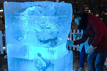 Ein Mann in einer leuchtend roten Jacke hackt energisch in einen massiven, blau leuchtenden Eisblock, der die schwach beleuchtete, urbane Nachtszene dominiert. Der strukturierte Eisblock, der auf einem dunklen Sockel steht und von einem weißen Lattenzaun umgeben ist, wird von dem Mann bearbeitet. Ein Klick auf das Bild führt zu einer größeren Ansicht.