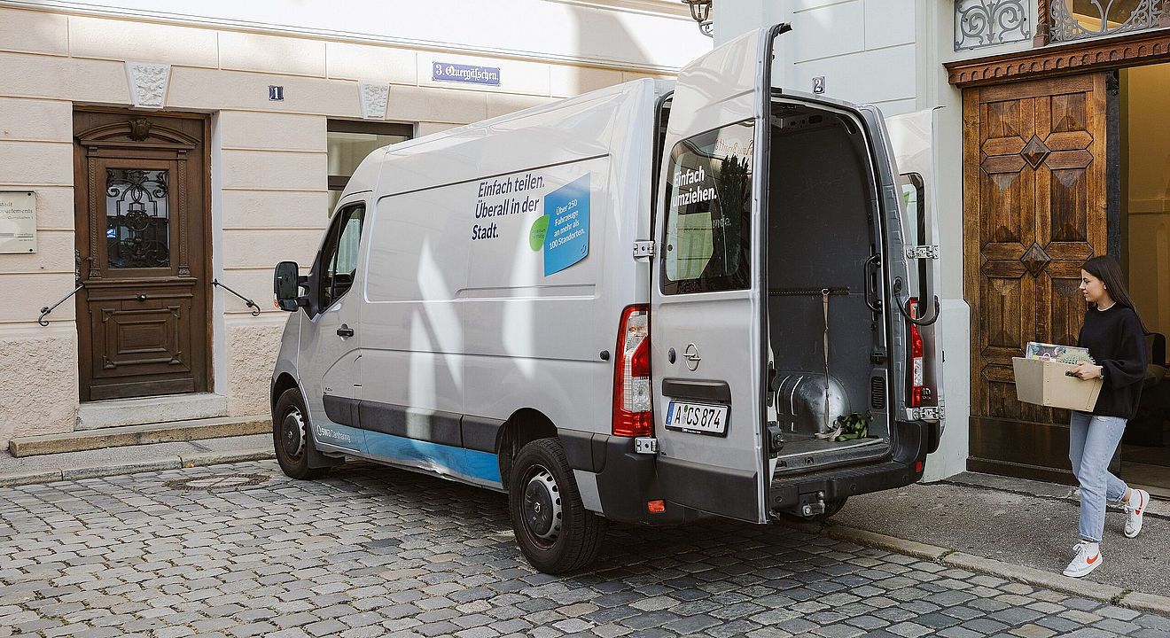 Frau trägt einen vollen Karton in einen geöffenten swa Carsharing Transporter. Ein Klick sorgt für eine Großansicht des Bildes.