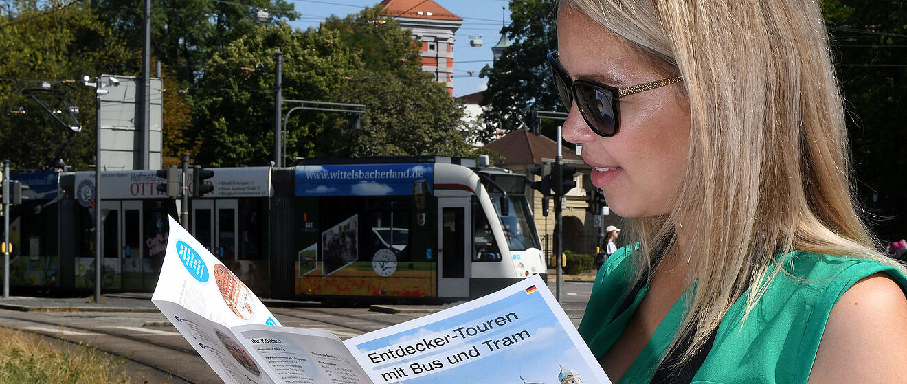 Frau mit Sonnenbrille schaut in ein Programmheft der Entdeckertour. Im Hintergrund ist eine Straßenbahn zu sehen.