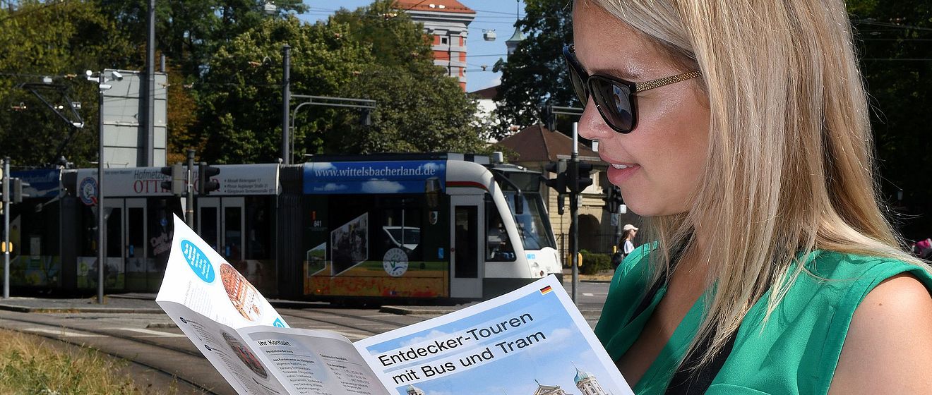 Frau mit Sonnenbrille schaut in ein Programmheft der Entdeckertour. Im Hintergrund ist eine Straßenbahn zu sehen.