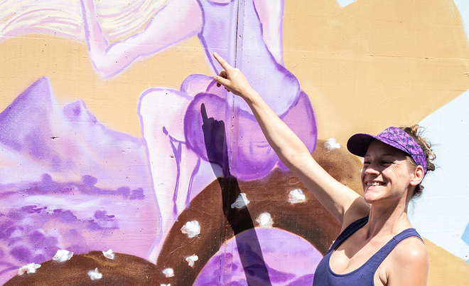 Im VorSophie Te guckt in einem dunkelblauen Kleid und einem lila Visor in die Kamera, während sie mit dem rechten Arm auf ein großes Wandgemälde hinter ihr zeigt.