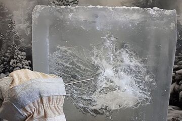 Ein mit Arbeitshandschuhen ausgestatteter Arm verwendet einen Eispickel, um in einen großen Eisblock zu hacken. Ein Klick auf das Bild führt zu einer größeren Ansicht.