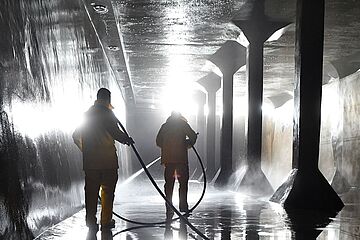 Zwei Personen in gelben Anzügen laufen mit einem Schlauch durch einen Hochbehälter. - Ein Klick auf das Bild führt zur vergrößerten Fotoansicht.