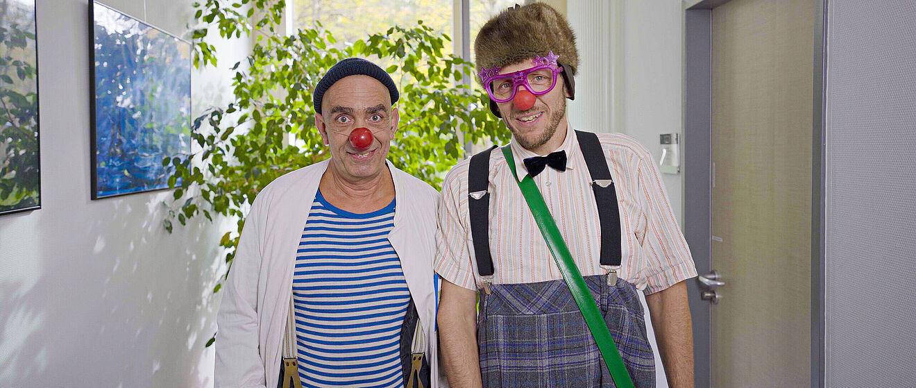 Zwei Männer der KlinikClowns in einem Clownskostüm stehen in einem Flur eines Krankenhauses vor einer Pflanze.