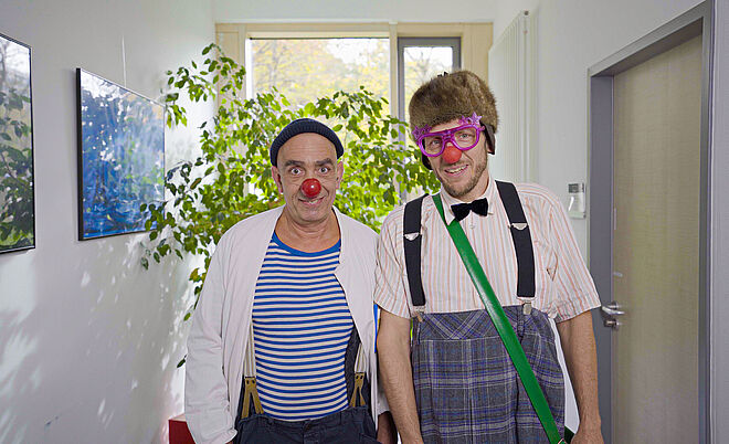 Zwei Männer der KlinikClowns in einem Clownskostüm stehen in einem Flur eines Krankenhauses vor einer Pflanze.