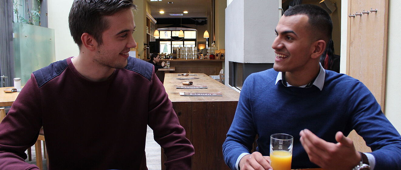 Zwei junge Männer führen ein angeregtes Gespräch in einem Cafe. Im Hintergrund befinden sich weitere Einrichtungen des Lokals.
