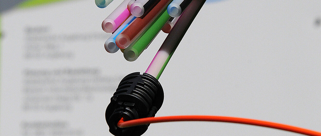 Dünne, farbige Glasfaserkabel treten aus einem schwarzen Stecker hervor und befinden sich vor einem Dokument mit dem Logo der swa.