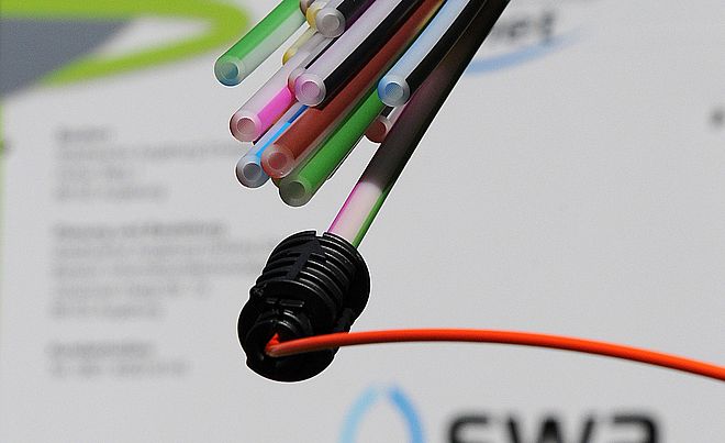 Dünne, farbige Glasfaserkabel treten aus einem schwarzen Stecker hervor und befinden sich vor einem Dokument mit dem Logo der swa.