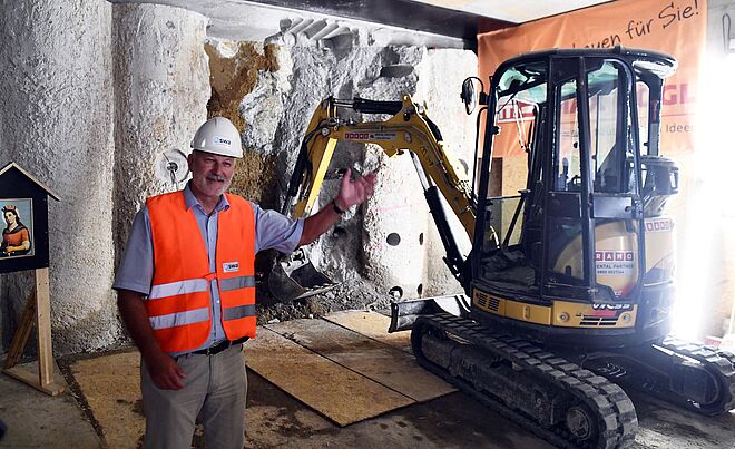 Mann trägt Schutzkleidung mit swa Branding und zeigt auf einen Bagger der ein Loch in der Wand gräbt.