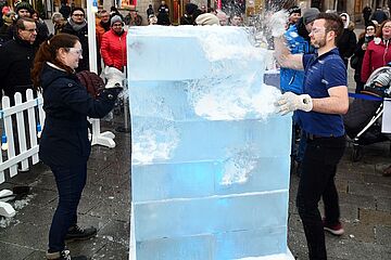 Eine Gruppe von Menschen hat sich um einen großen, beleuchteten Eisblock versammelt, der auf einem belebten, nassen Platz steht. Zwei Teilnehmer, bekleidet mit schwarzen und blauen Jacken sowie Schutzbrillen, hacken mit Eispickeln auf das Eis ein, wobei Eisstücke durch die Luft spritzen. Ein Klick auf das Bild führt zu einer größeren Ansicht.