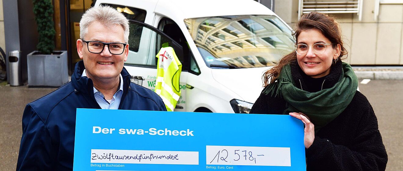 Ein Mann und eine Frau halten einen großen blauen swa-Spendenscheck im Wert von 12.578 Euro für SKM Augsburg