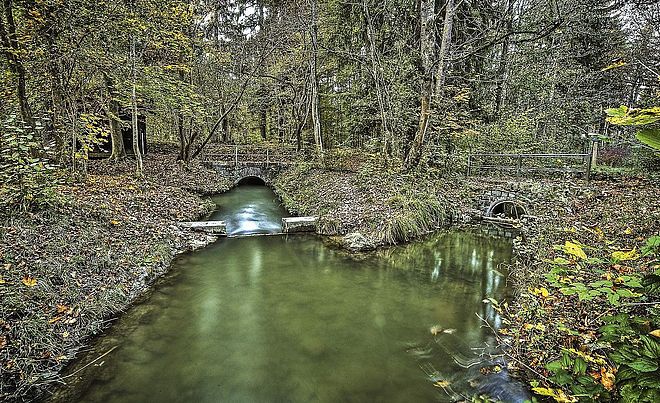 Idyllische, im Wald gelegene Wasserkreuzung, in welche Mauerreste hineinragen. Im Hintergrund befinden sich zudem zwei Brücken über den Wasserläufen.