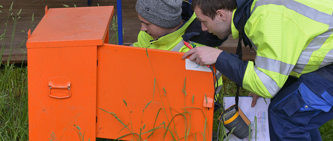 Zwei Männer in gelben Warnwesten blicken in einen orangen (Strom-)Kasten.