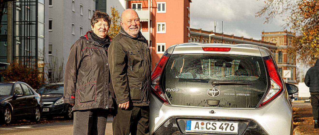 Ein Mann und eine Frau stehen hinter einem Carsharing Auto