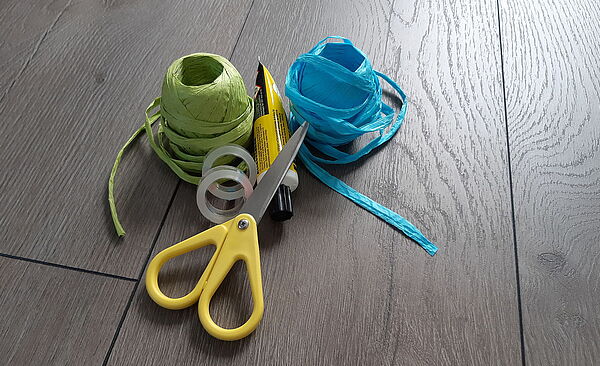 Eine Schere, ein Kleber, zwei Klebebänder und zwei Geschenkbänder liegen auf einem Tisch 