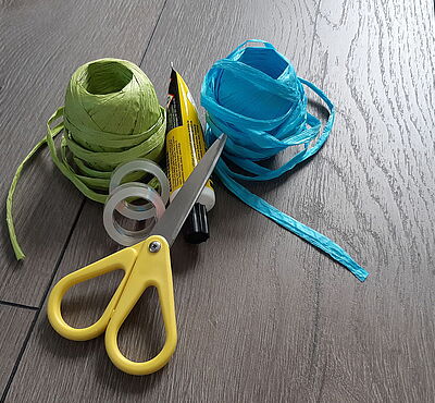 Eine Schere, ein Kleber, zwei Klebebänder und zwei Geschenkbänder liegen auf einem Tisch